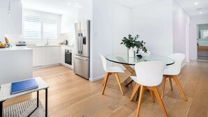 A modern kitchen with white cabinets, a stainless steel refrigerator, and a dining area with a glass table and white chairs. - Top Floor Apartment Close To Bondi Beach (Bondi)
