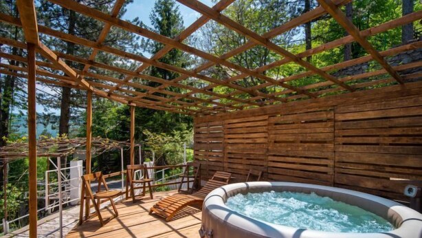 A hot tub under a wooden pergola with chairs, surrounded by a wooden deck and a stone wall. - Vacation home Nina by Interhome (Fužine)