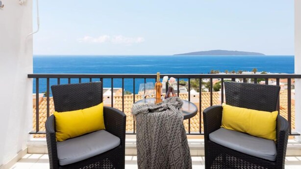 Un balcone con due poltrone in vimini, ognuna con un cuscino giallo, affaccia su una vista costiera che include il mare e un'isola in lontananza.