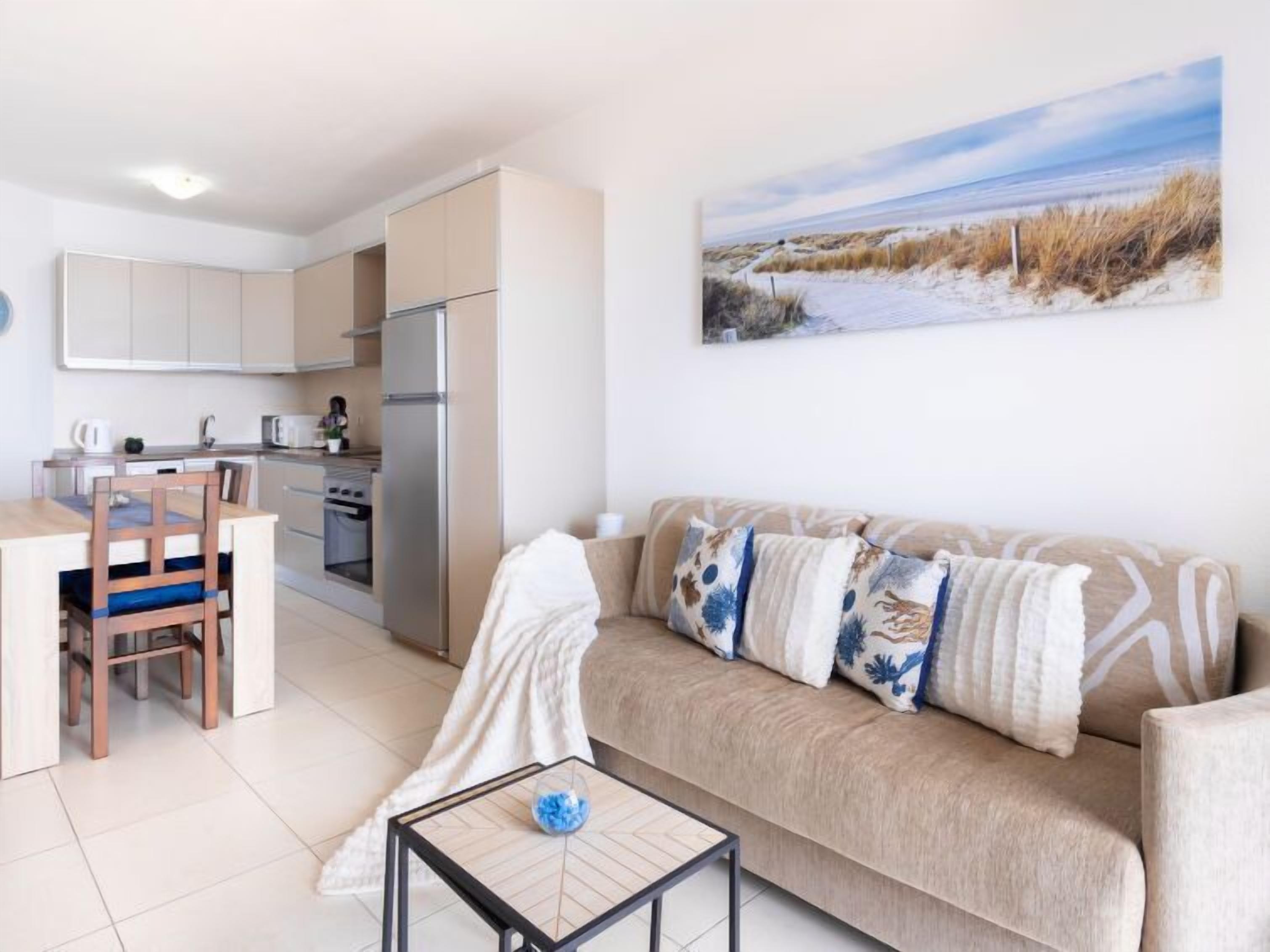 A modern apartment with a kitchenette, a sofa with patterned cushions, and a painting of a beach scene.