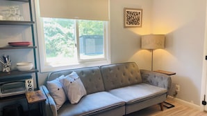 A living room with a grey sofa, a wooden side table, a lamp, and a window with a view of greenery. - Newly Renovated - Suite Springs (Yellow Springs)