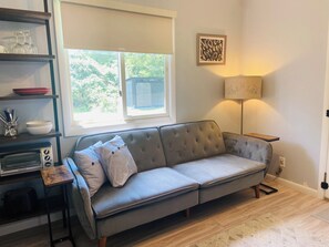 A living room with a grey sofa, a wooden side table, a lamp, and a window with a view of greenery.