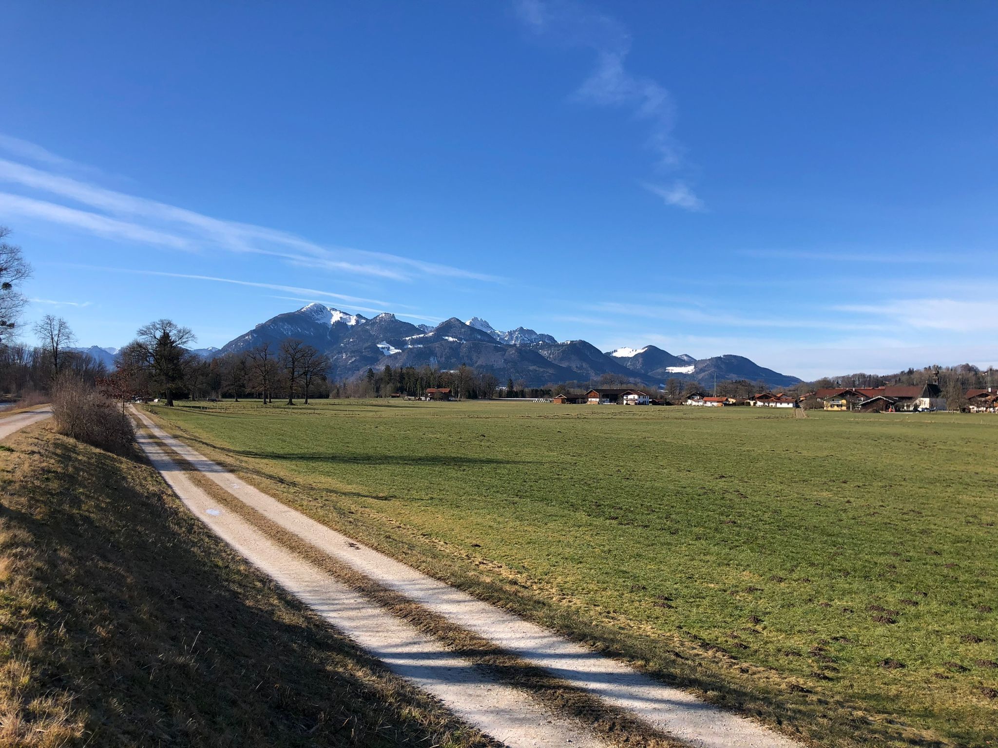 Paesaggio rurale con un sentiero sterrato che conduce a un villaggio, circondato da montagne e campi aperti.