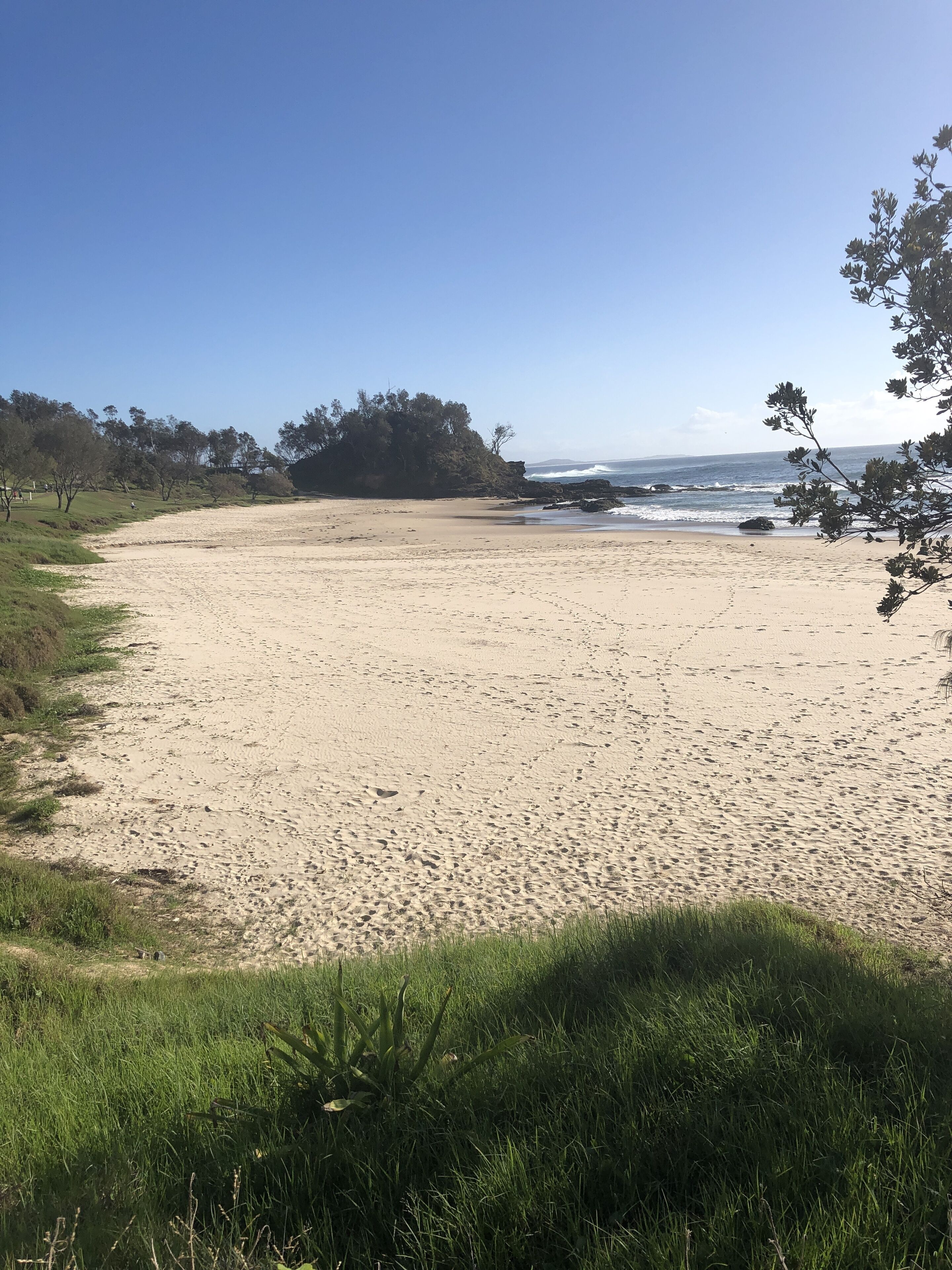 Een zandstrand met een grasveld op de voorgrond en een rotsformatie in de verte.