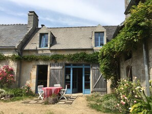 Een rustiek stenen huis met een schuin dak, een houten deur en een kleine tuin met bloemen en een tafel met stoelen.