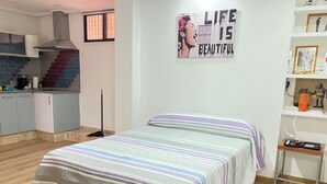 Quarto com cama, mesa de cabeceira, parede com pôster e área de cozinha ao fundo.