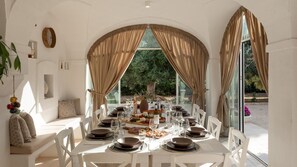 A dining area with a table set for six, large windows with curtains, and a view of greenery outside.