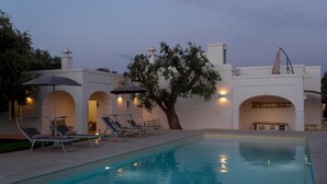 A pool area with lounge chairs and umbrellas, a tree, and a building with arches and a chimney. - La Torre di Monsignore 12 2 (Fasano)