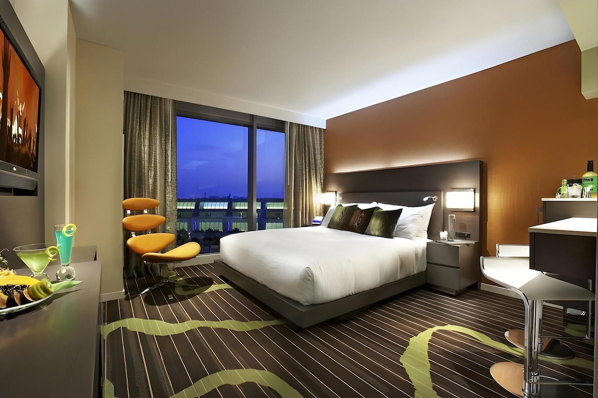 Een hotelkamer met een groot bed, een bureau met stoel, een minibar en uitzicht op de stad in de schemering.