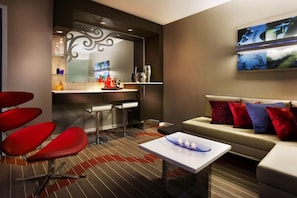 Une chambre d’hôtel moderne avec un coin bar, un canapé agrémenté de coussins rouges et bleus, et une table basse avec des bougies.