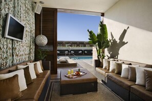 Un salón moderno con vista a la piscina, un televisor de pantalla plana y un comedor con una bandeja de frutas y bebidas.