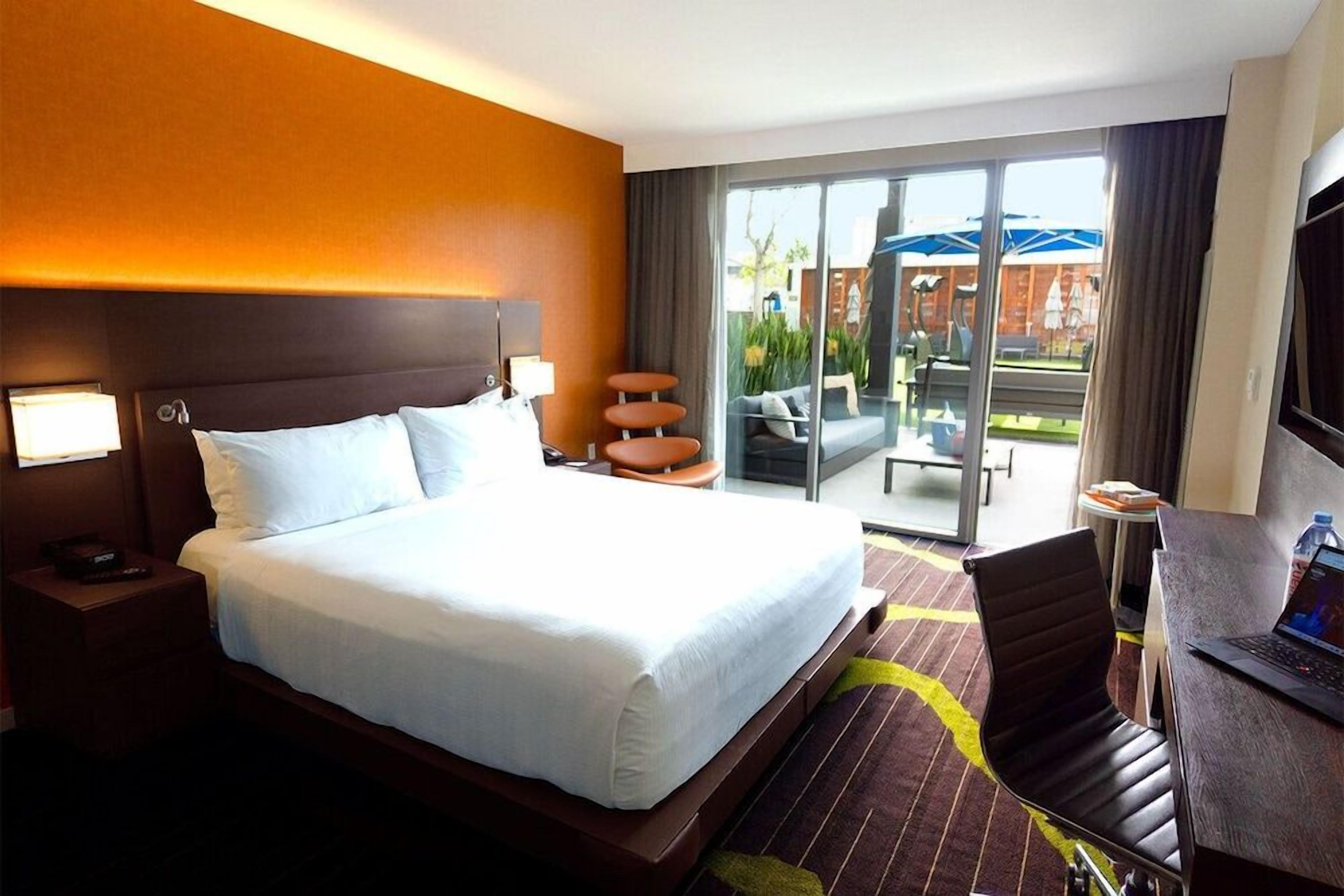 Habitación de hotel con una cama grande, un escritorio con una computadora portátil, un televisor y un balcón con mobiliario exterior.