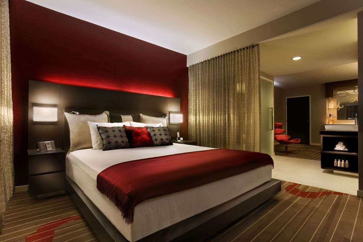 Una habitación de hotel moderna con una cama grande, lámparas de noche, una pared de acento roja y vistas a un área de salón.
