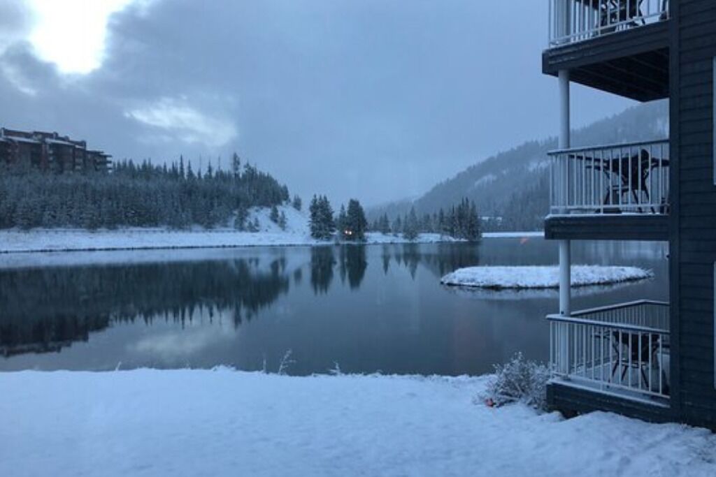 A snow-covered landscape with a calm lake, a forested hill, and a building with a balcony.