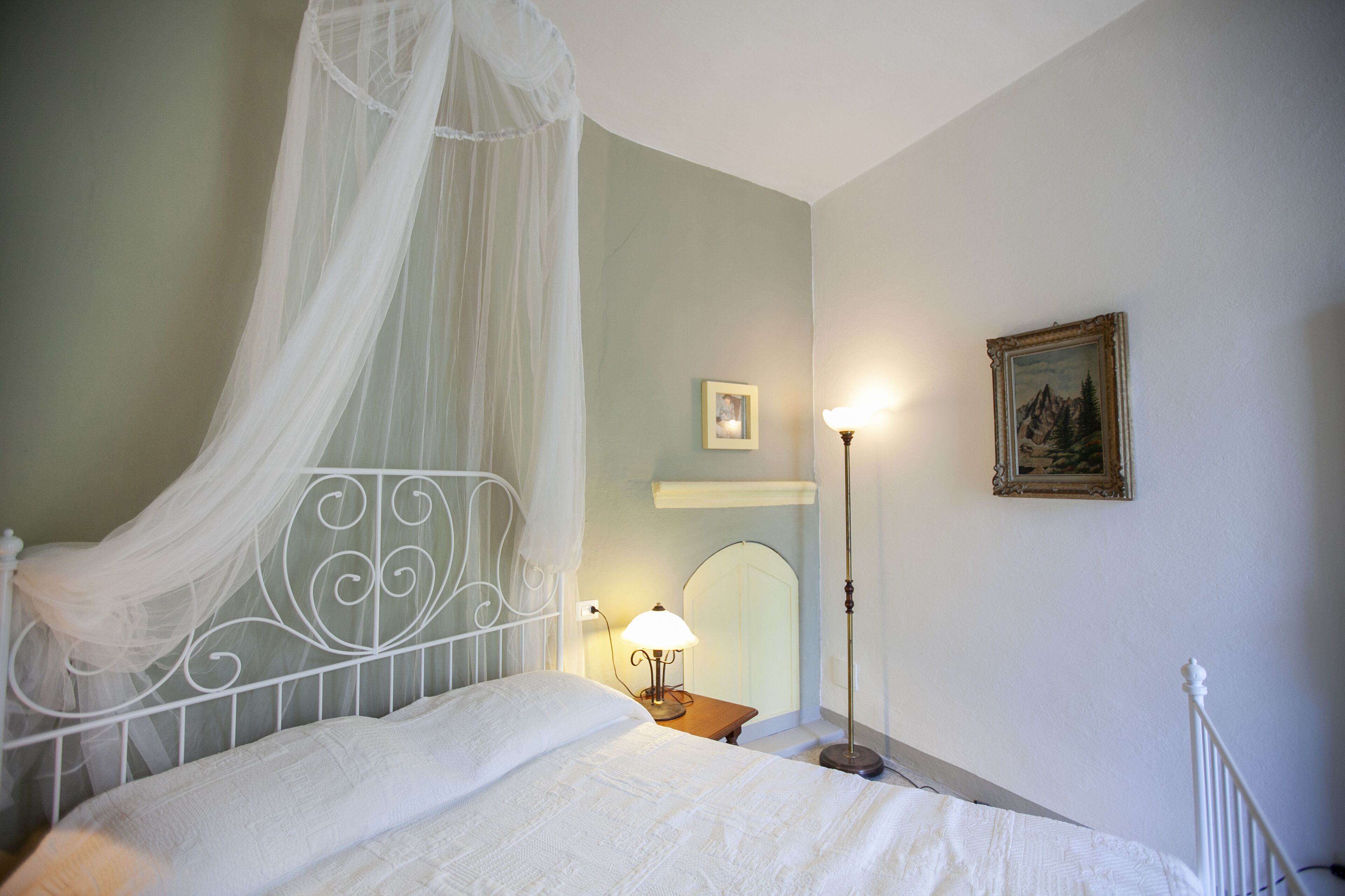 A bedroom with a four-poster bed, a bedside table with a lamp, a framed picture on the wall, and a mirror above the table.