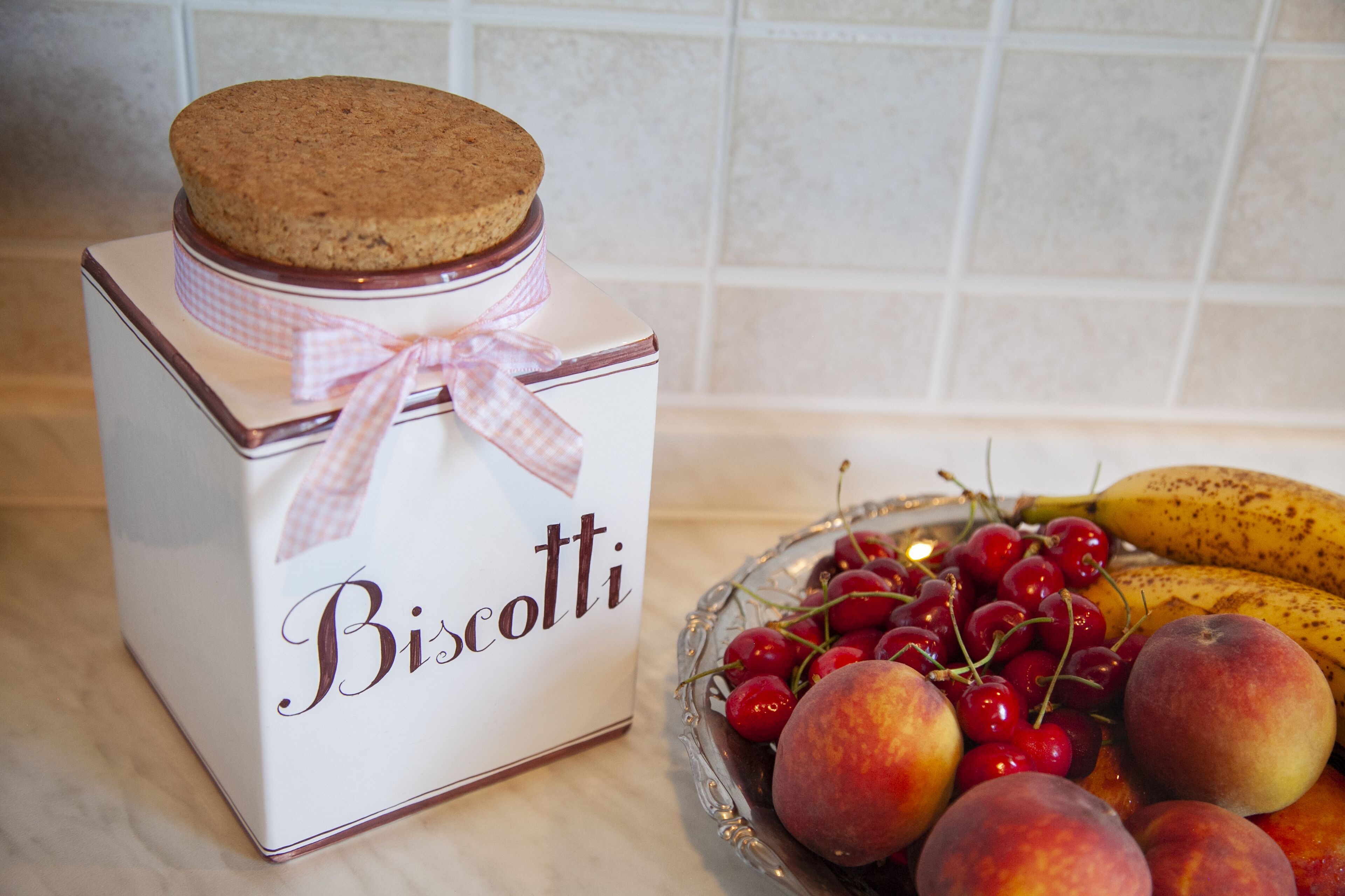 Eine Schachtel mit dem Aufdruck "Biscotti", geschmückt mit einer karierten Schleife, eine Glaskasse mit Kirschen, Pfirsichen und einer Banane.
