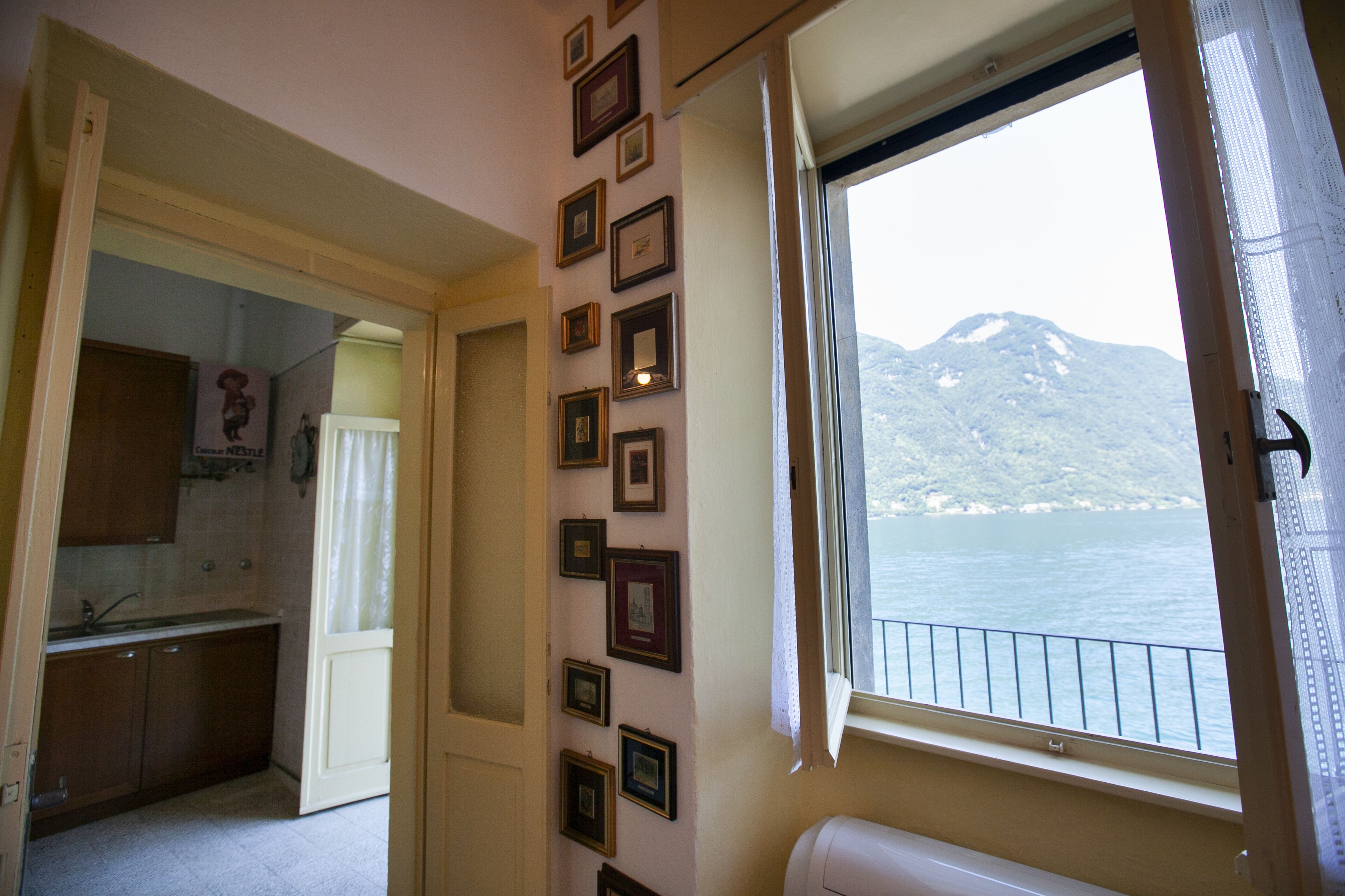A room with a window overlooking a body of water and mountains, a kitchen area, and a wall decorated with framed pictures.