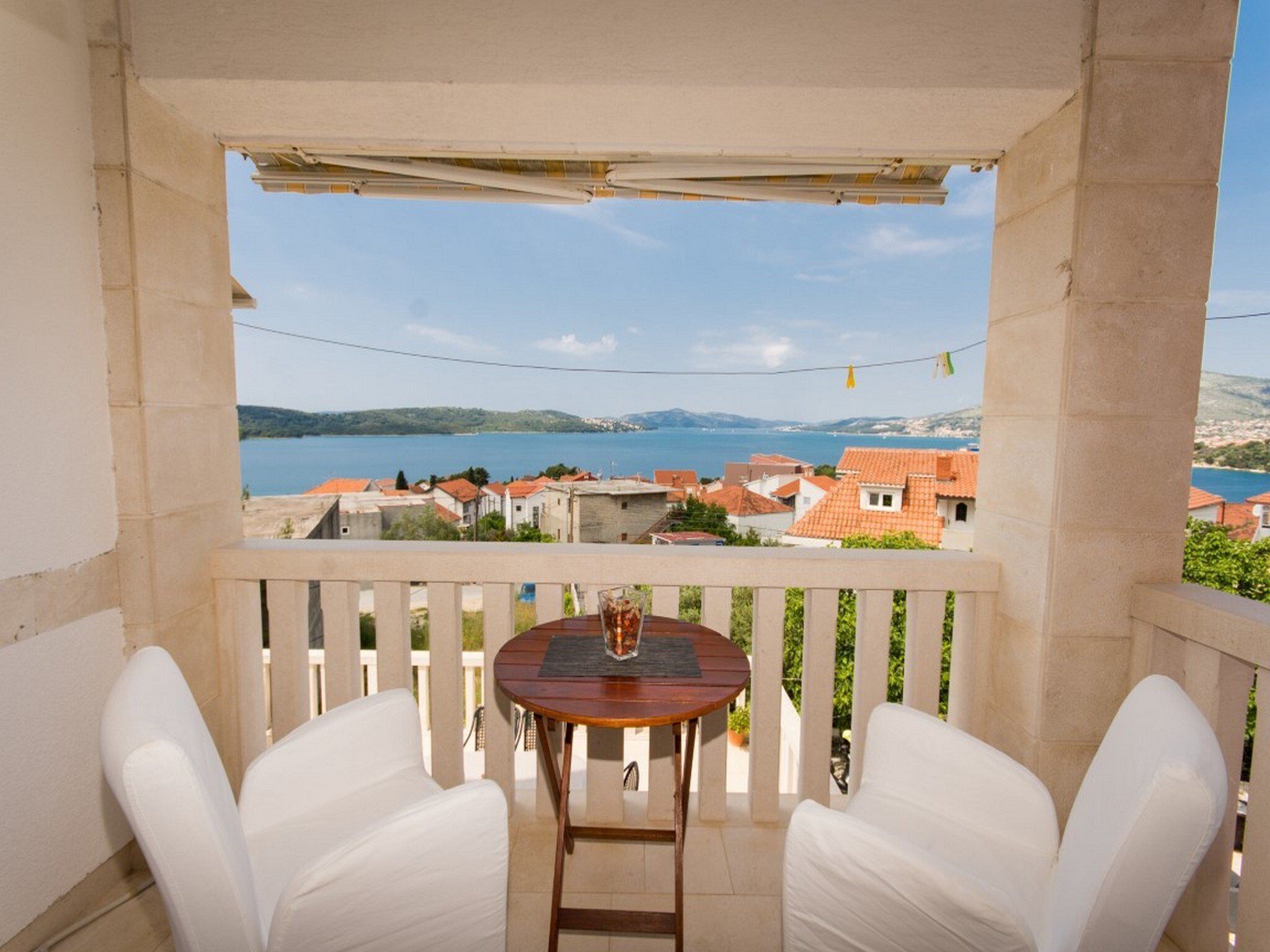 A balcony with a table and two chairs, overlooking a coastal town with red-roofed buildings and a body of water.