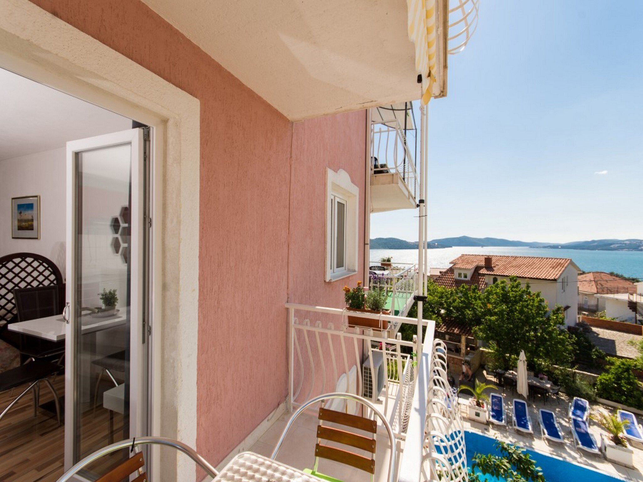 A balcony with a view of a lake and mountains, a dining area with a table and chairs, and a pool area with lounge chairs.