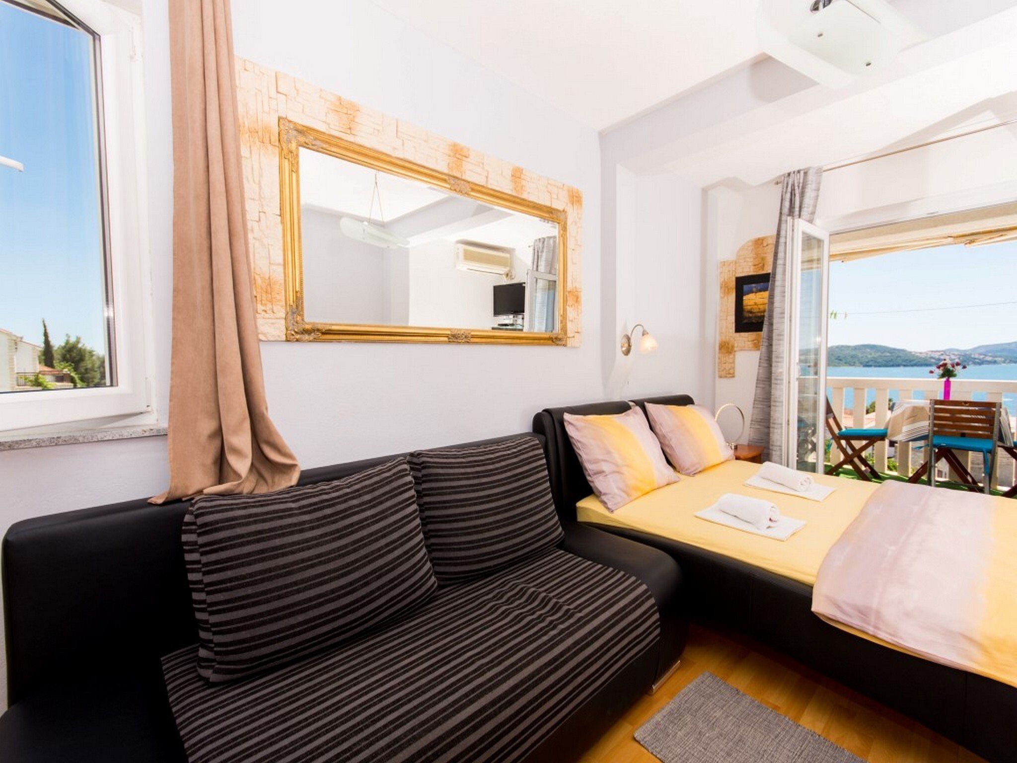 A modern bedroom with a large bed, a black sofa, a mirror, and a view of the sea.