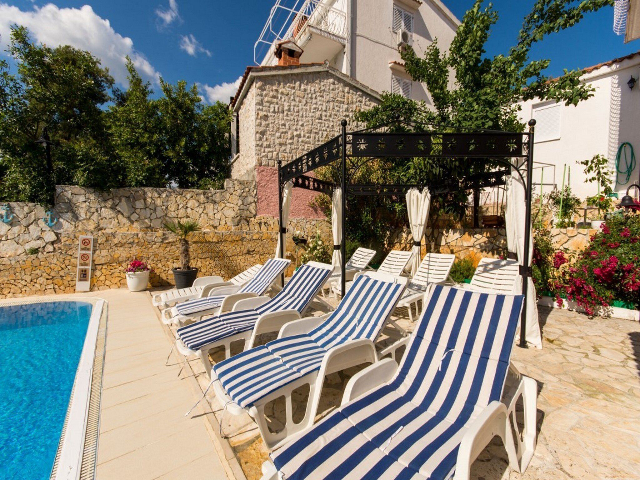 Un espace aménagé au bord de la piscine, avec des chaises longues à rayures, un mur en pierre et un bâtiment doté d’un balcon.