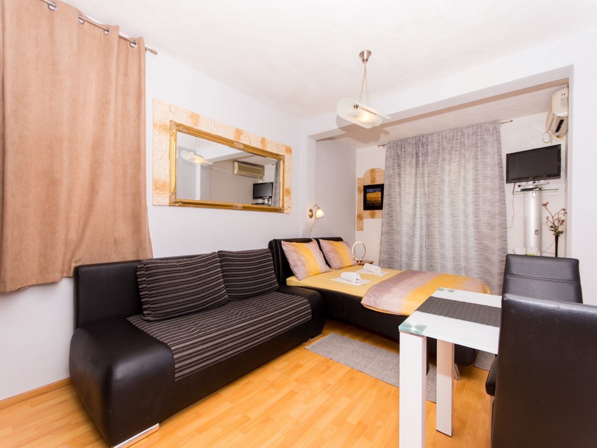 A modern hotel room with a black sofa, a small table, a bed with a yellow and orange blanket, a large mirror, a ceiling fan, and a TV.