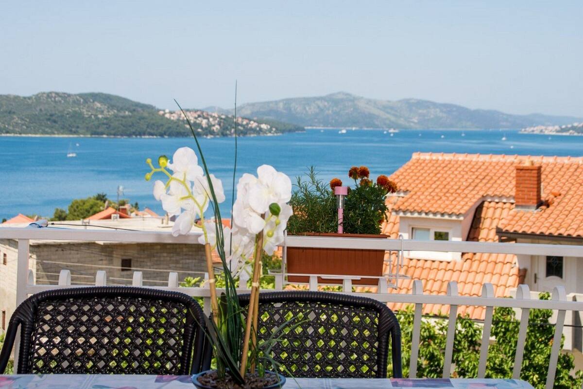 A terrace with wicker chairs and a table, overlooking a coastal view with houses and the sea.