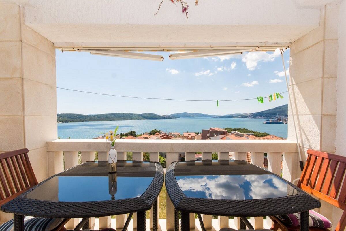 A balcony with two wicker chairs and a table, overlooking a lake and distant hills.