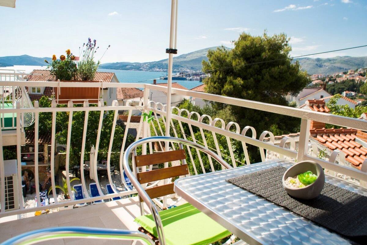 A balcony with a view of a lake and mountains, a table with a bowl, and a chair.