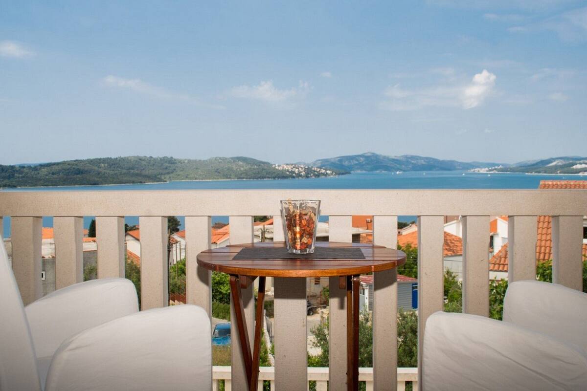 A balcony with a wooden table and two white chairs, overlooking a body of water and distant hills.