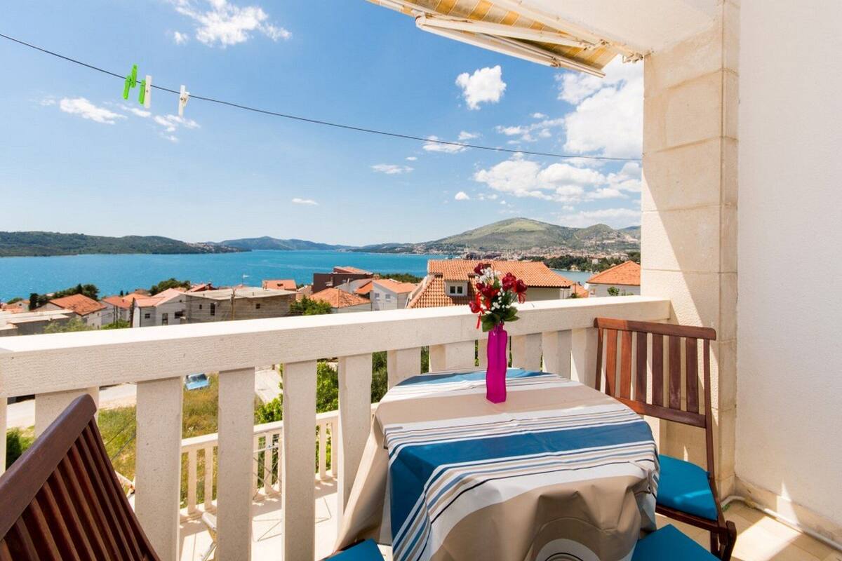 A balcony with a view of the sea, a table with a striped tablecloth, and a chair.