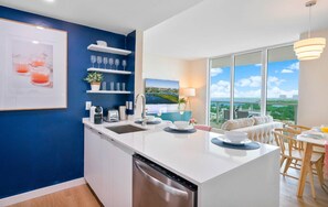 A modern kitchen with a dishwasher, a dining area with a table and chairs, and a large window offering a view of the city.