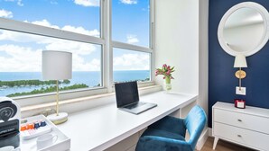 A room with a laptop on a white desk, a blue chair, a white dresser, a round mirror on the wall, and a view of the sea through large windows. - Peacock Park Steps Away | Modern Residence (Miami)