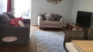 Een woonkamer met een donkergrijze bank, een lichtgrijze bank, een houten salontafel, een blauw pluche vloerkleed en een televisie op een houten dressoir.