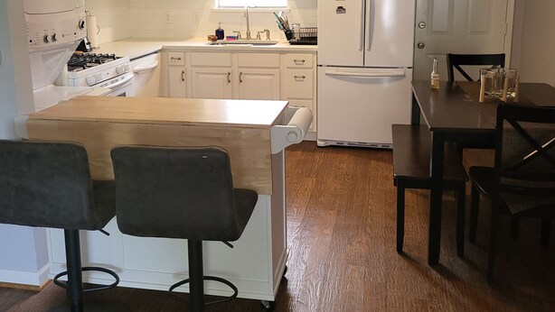 Een moderne keuken met witte kasten, een centraal eiland met barstoelen, een koelkast en een eettafel met stoelen.