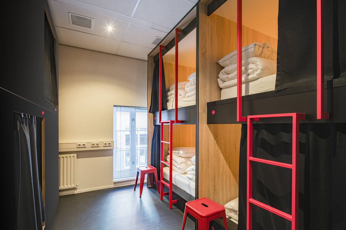 A hotel room with a bunk bed, a radiator, a window with blinds, and a red stool.