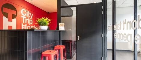 A modern hotel lobby with a red accent wall, a reception desk, and two red bar stools.