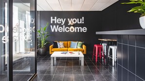 A modern reception area with a black wall, a yellow sofa, a white coffee table, and a red stool. - City Trip Hostels (Purmerend)