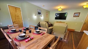 A dining area with a wooden table set for four, a sofa, and a television mounted on the wall. - {Forget-Me-Not Inn} Alaskan Vacation Rental (Bear Suite) (Sitka)