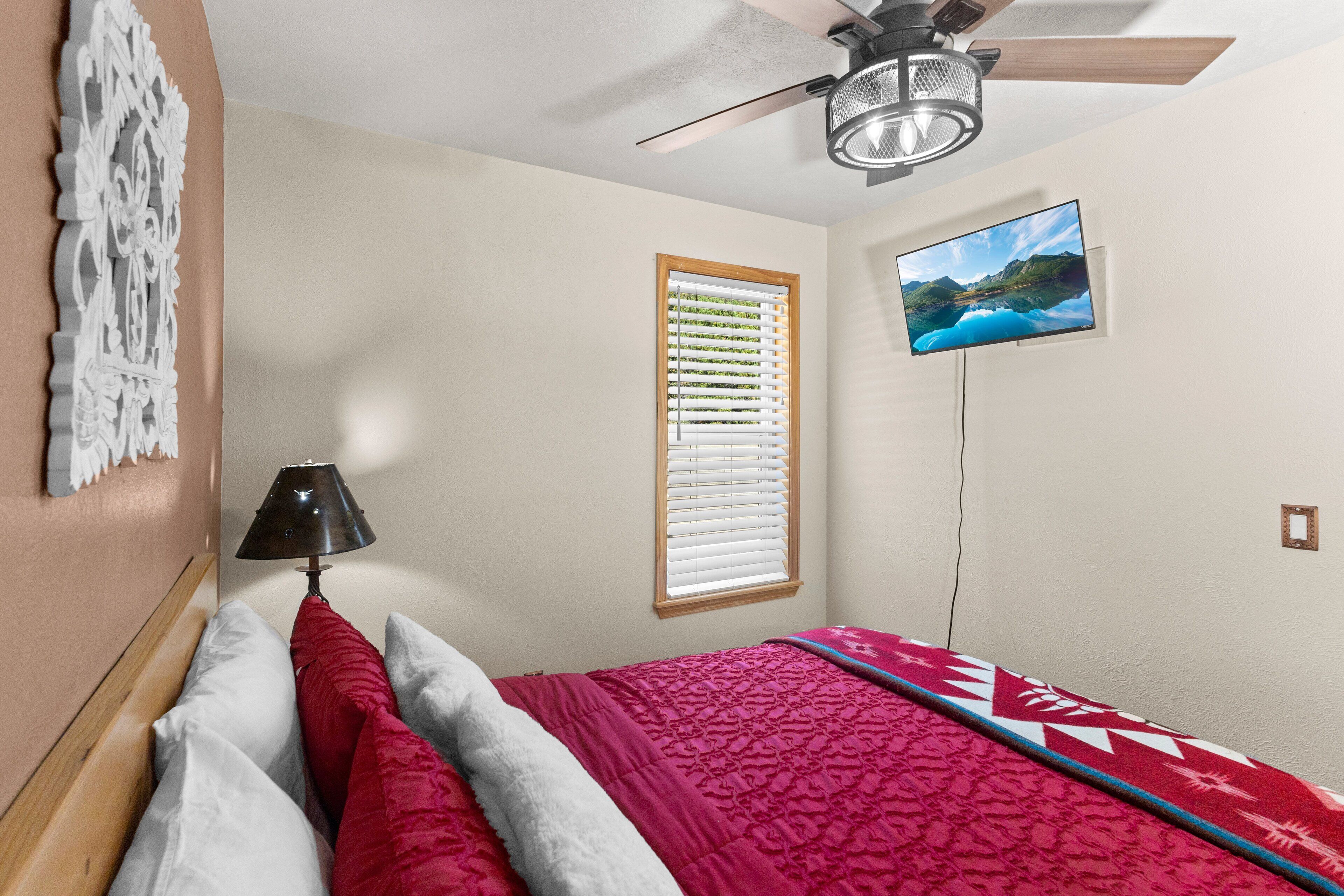 A bedroom with a bed, a ceiling fan, a TV mounted on the wall, and a window with blinds.
