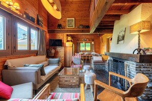A cozy log cabin interior with a fireplace, wooden furniture, and large windows offering a view of the outdoors.