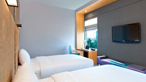 A hotel room with two beds, a desk with a computer, a TV mounted on the wall, and a window with a view of buildings. - Hotel Halez (Chennai)