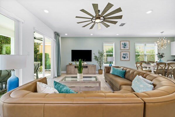 A modern living room with a large brown sectional sofa, a glass coffee table, a ceiling fan, and a flat-screen TV.