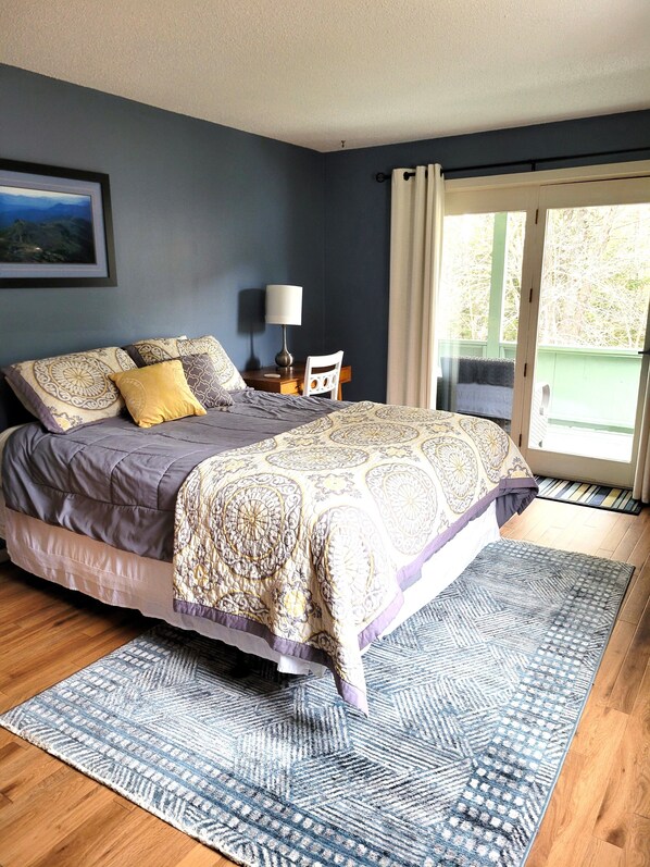 A bedroom with a bed, a nightstand, a chair, a lamp, and a view of the outdoors through a window.