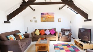 A spacious living room with a high ceiling, exposed wooden beams, a large window, a sofa set, a coffee table, a television, and a colorful rug. - The Corn Tallet (Bideford)
