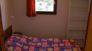 Un dormitorio con una cama cubierta con un colcha colorida, una ventana con cortina y un estante blanco.