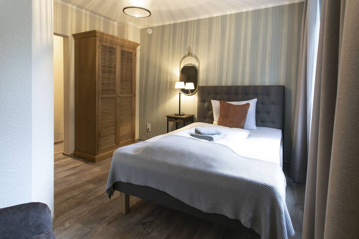 A hotel room with a bed, a nightstand with a lamp, a wardrobe, and a mirror on the wall.