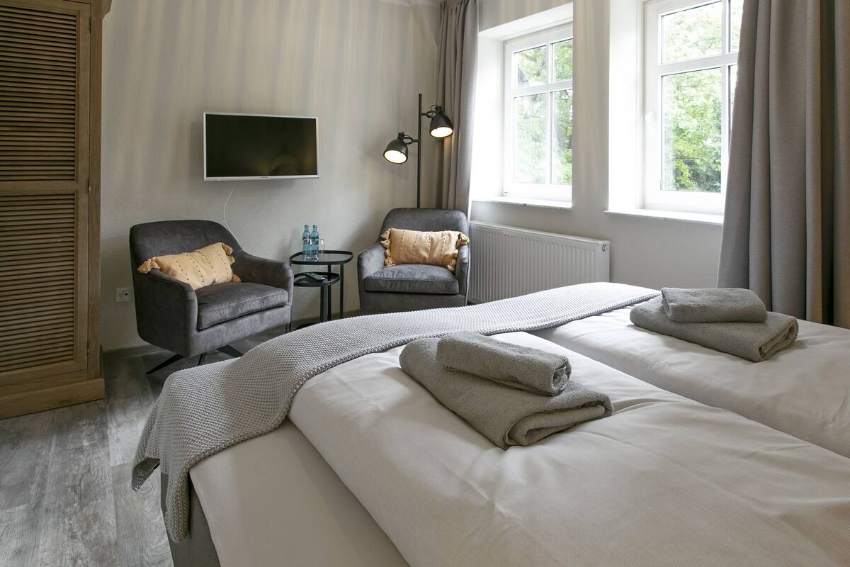 A hotel room with two beds, a TV, a chair, a small table, and a window with curtains.
