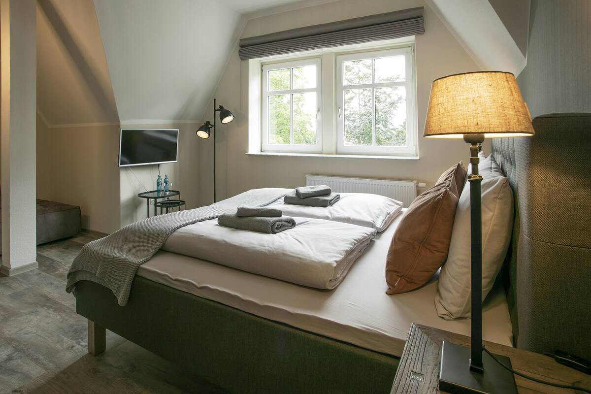 A neatly made bed with pillows and a blanket, a bedside lamp, a flat-screen TV, and a window with daylight.