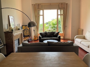 A living room with a fireplace, a sofa, a dining table, and a view of greenery outside. - Beautiful Haussmann apartment in the Golden Square (Marseille)
